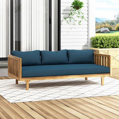 The Claremont 3 Seater Daybed, crafted from teak acacia wood with water-resistant blue cushions, is placed on a white patterned rug atop a wooden deck, set against glass doors and lush outdoor greenery.