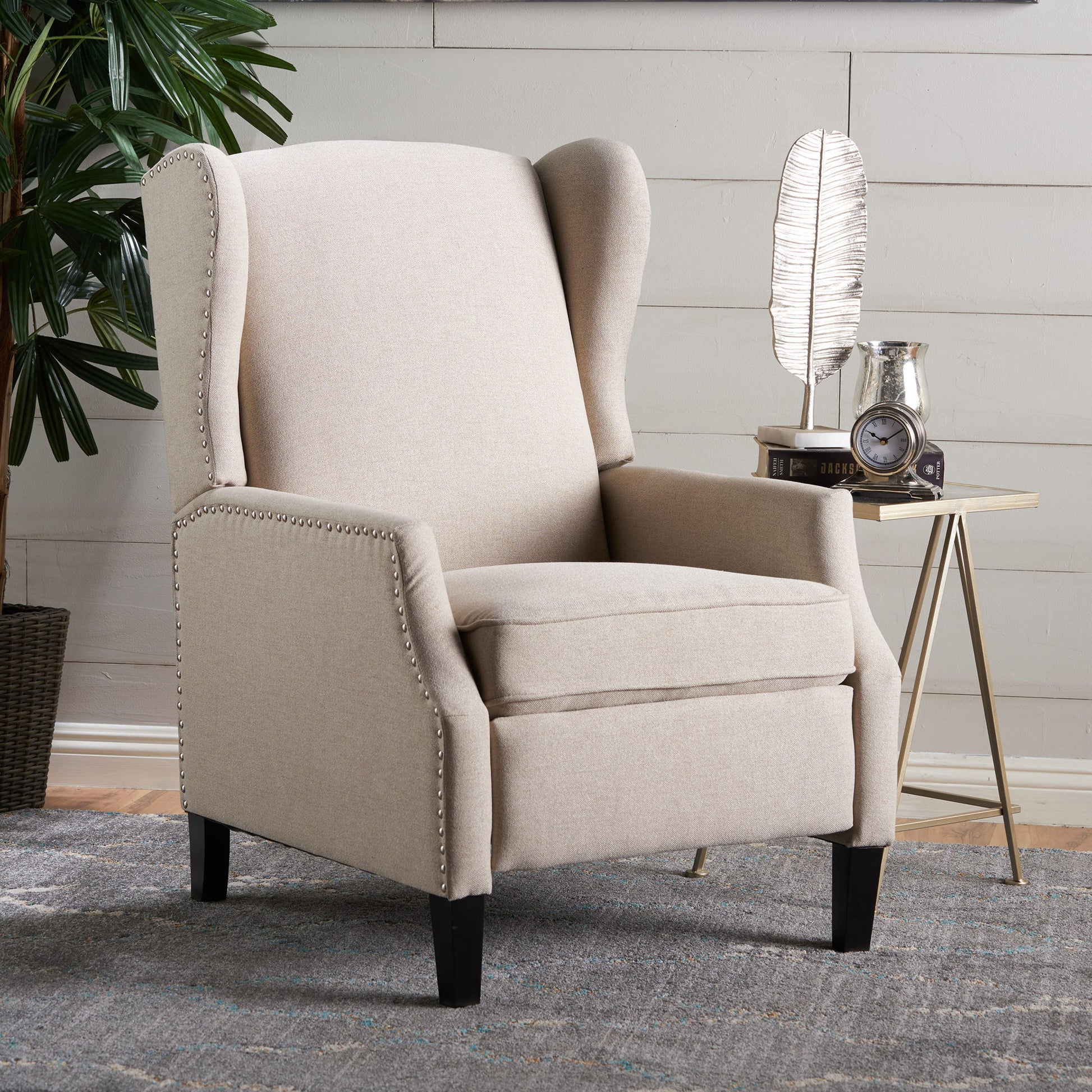 The Westeros Wing Back Fabric Upholstered Recliner in beige with nailhead trim is set on a patterned rug, next to a side table featuring a feather-shaped decor piece, clock, and pitcher. A leafy plant appears in the background.