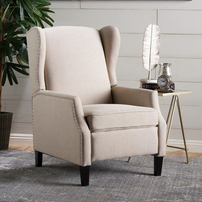 The Westeros Wing Back Fabric Upholstered Recliner in beige with nailhead trim is set on a patterned rug, next to a side table featuring a feather-shaped decor piece, clock, and pitcher. A leafy plant appears in the background.