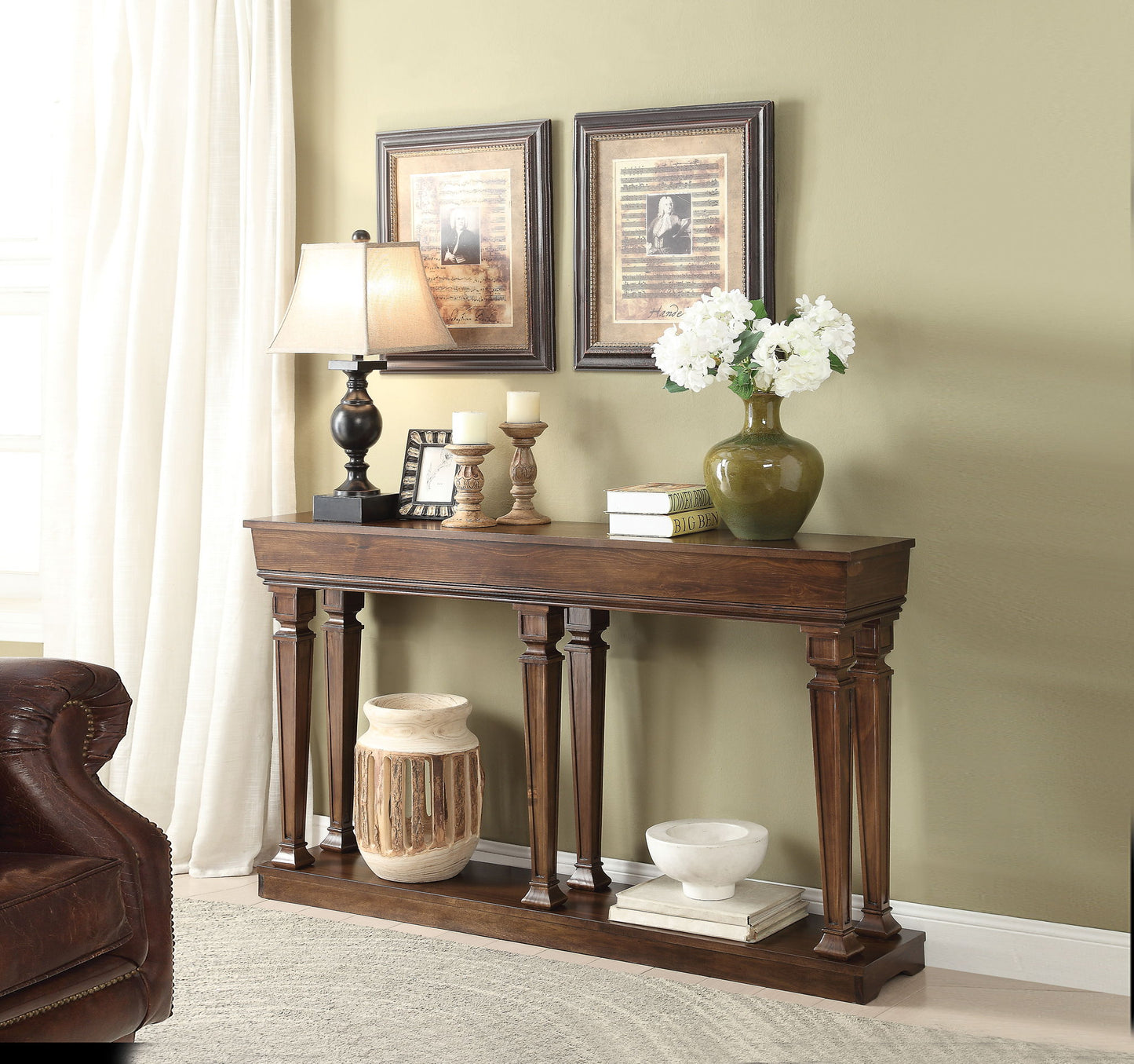 The Garrison - Wooden Console Table features decorative columns and displays a lamp, candlesticks, framed photos, books, and a green vase with white flowers. Beneath are a large ceramic jar and a white bowl. Two framed pictures hang above.