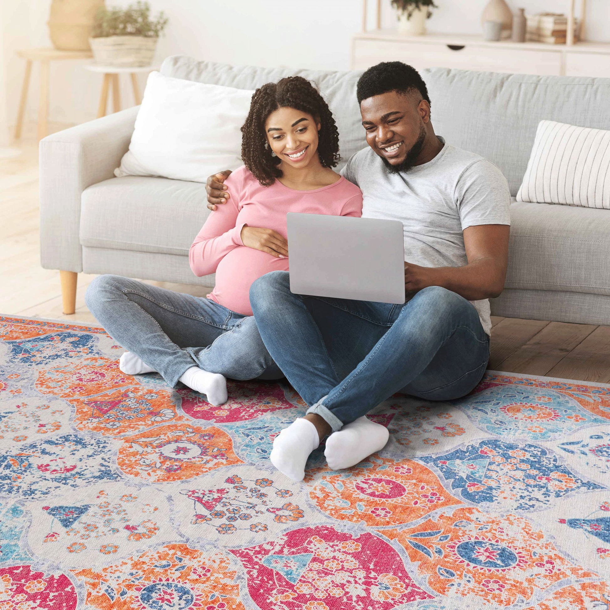 A pregnant woman and a man sit on the Maval - Stunning Area Rug, leaning against a gray sofa. Smiling and relaxed, they enjoy time together looking at a laptop in their cozy living room.