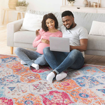 A pregnant woman and a man sit on the Maval - Stunning Area Rug, leaning against a gray sofa. Smiling and relaxed, they enjoy time together looking at a laptop in their cozy living room.