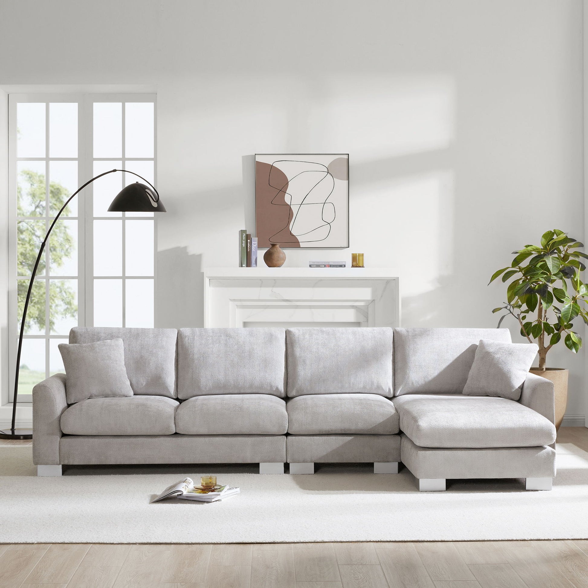 A modern living room featuring the Modern Oversized Sectional Sofa, an L-shaped luxury couch set with 2 free pillows and chaise, is accented by a floor lamp, green potted plant, abstract art above a white mantel, and sunlight through large windows.