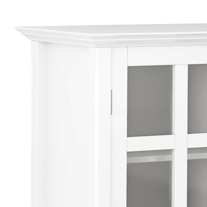 Close-up of the corner of the Acadian - Medium Storage Cabinet, Handcrafted. It features white wood, glass panel doors with decorative top trim, and shelves visible through the glass panels.