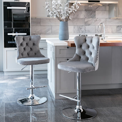 Two Swivel Velvet Barstools with tufted backs and chrome bases are placed beside a modern kitchen island with light cabinetry and a tiled backsplash. A vase of white flowers decorates the island. Set includes 2 adjustable upholstered bar stools.