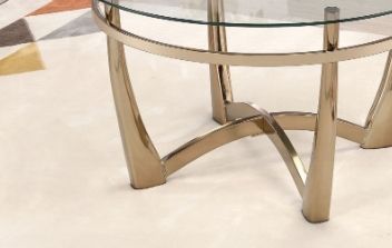 A close-up of the Orlando - Clear Glass Coffee Table - Champagne, featuring a round glass top and metallic curved legs, placed on a light floor with part of a geometric-patterned carpet in the background.