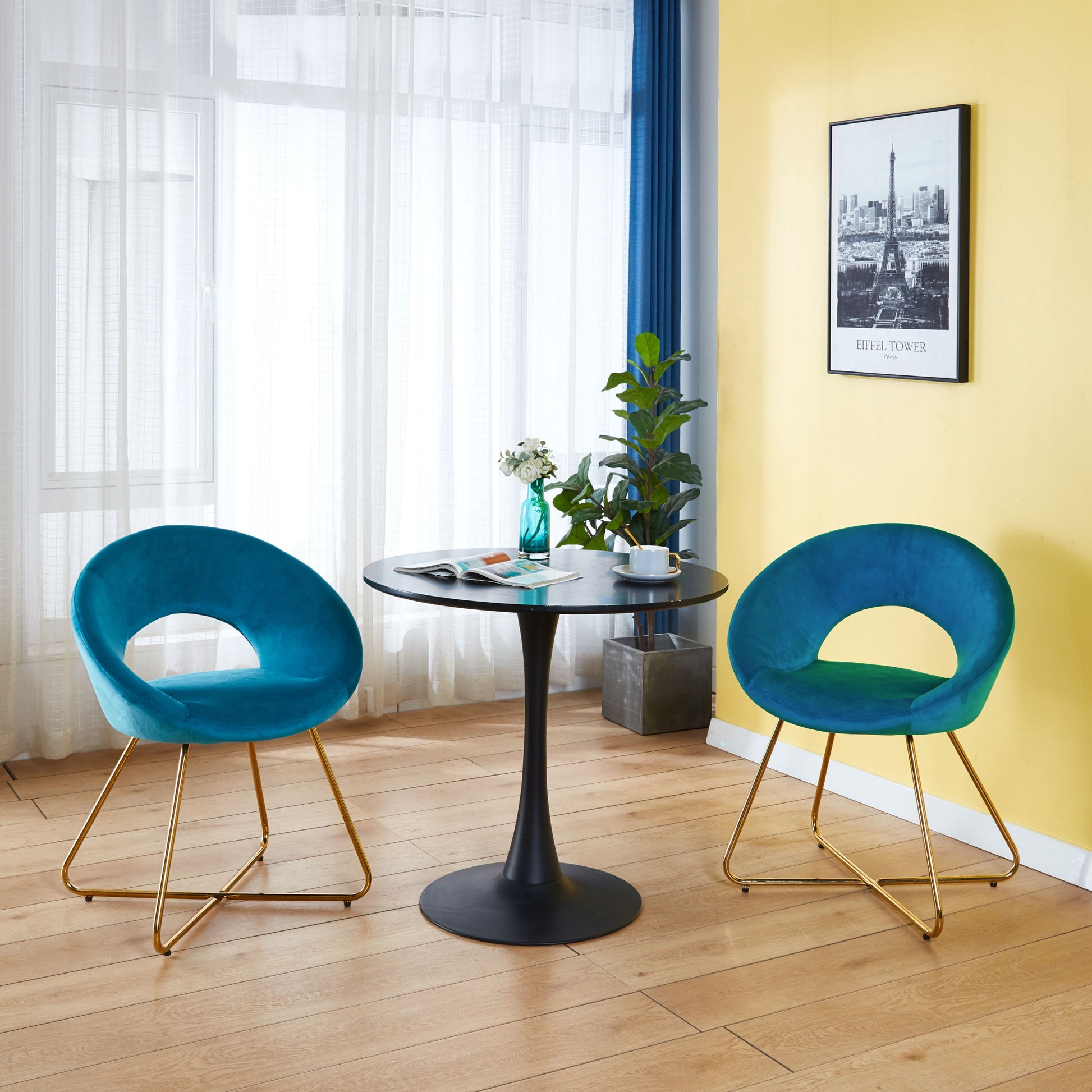 A modern room with two Modern Accent Velvet Lounge Chairs, a round black table with a vase and cup, a potted plant, wooden floor, yellow wall, and framed Eiffel Tower art. Large windows with sheer curtains brighten the space.