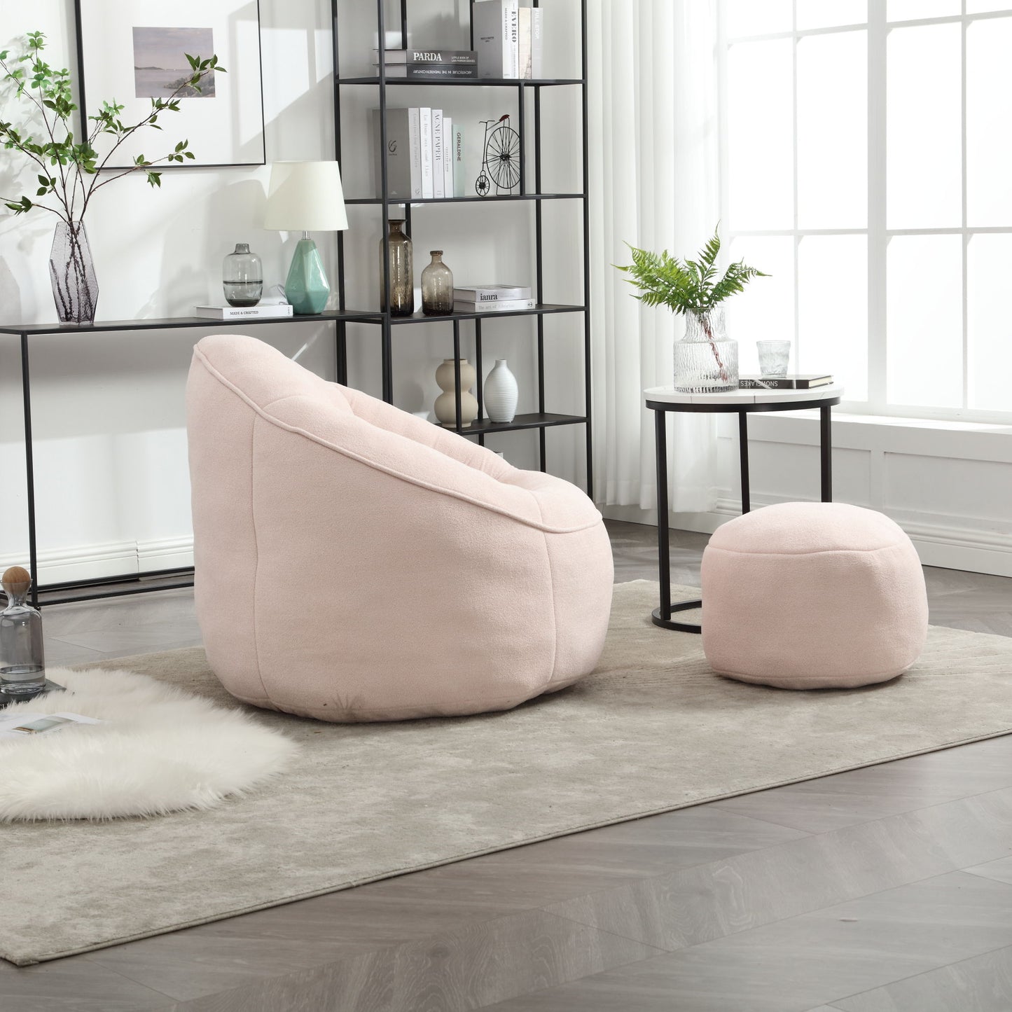 A cozy living room showcases the Bedding Bean Bag Sofa Chair High Pressure Foam with Footrest in pale pink on a gray rug, beside a side table with a vase, near a large window and black shelves holding decor and books.