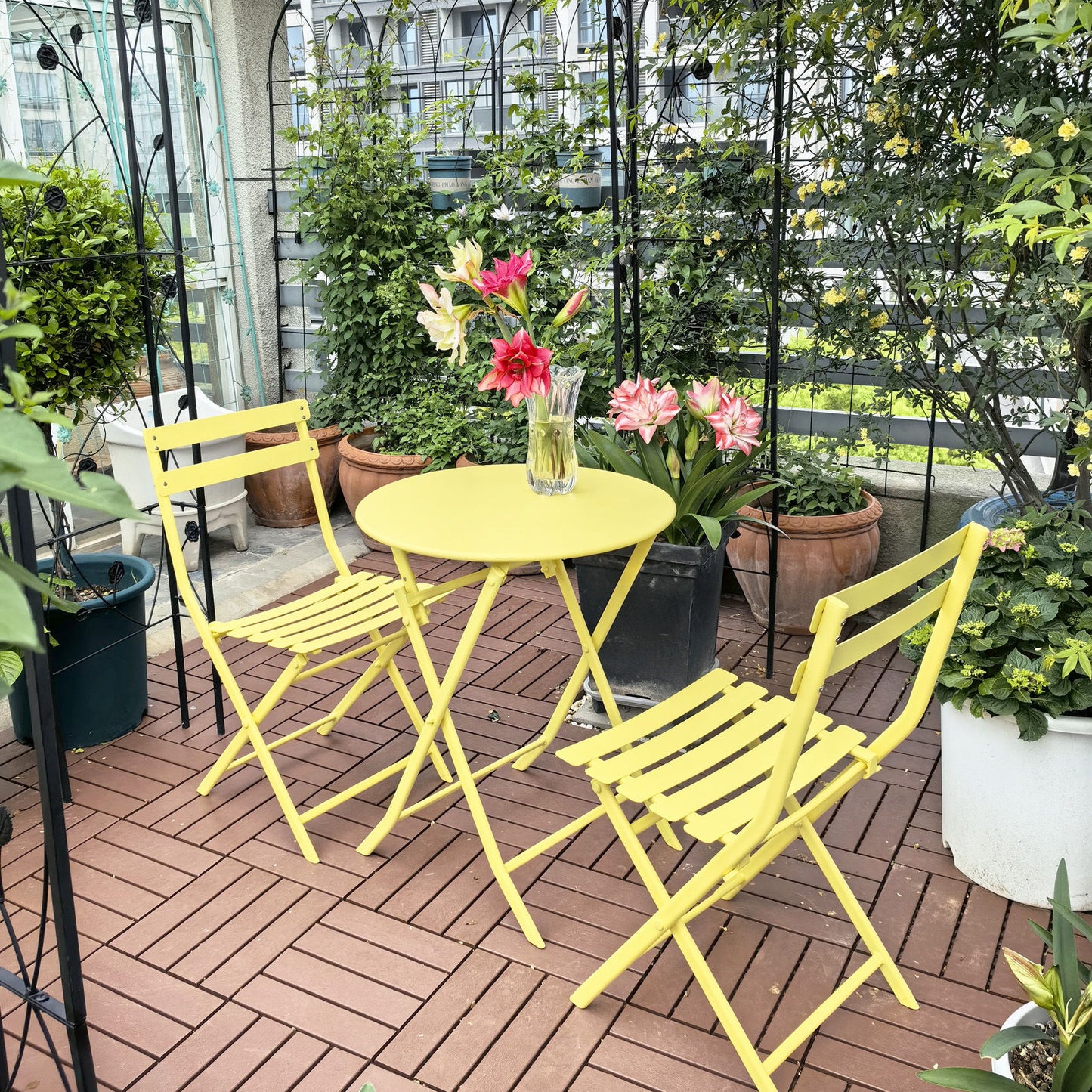 The 3 Piece Patio Bistro Set of Foldable Round Table and Chairs, in bright yellow, sits on a wooden deck surrounded by potted plants. A vase with colorful flowers on the table adds cheer to this cozy garden retreat.