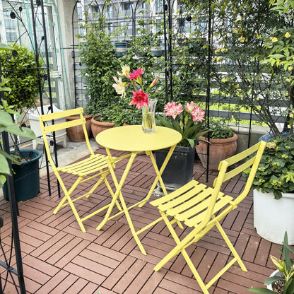 The 3 Piece Patio Bistro Set of Foldable Round Table and Chairs, in bright yellow, sits on a wooden deck surrounded by potted plants. A vase with colorful flowers on the table adds cheer to this cozy garden retreat.