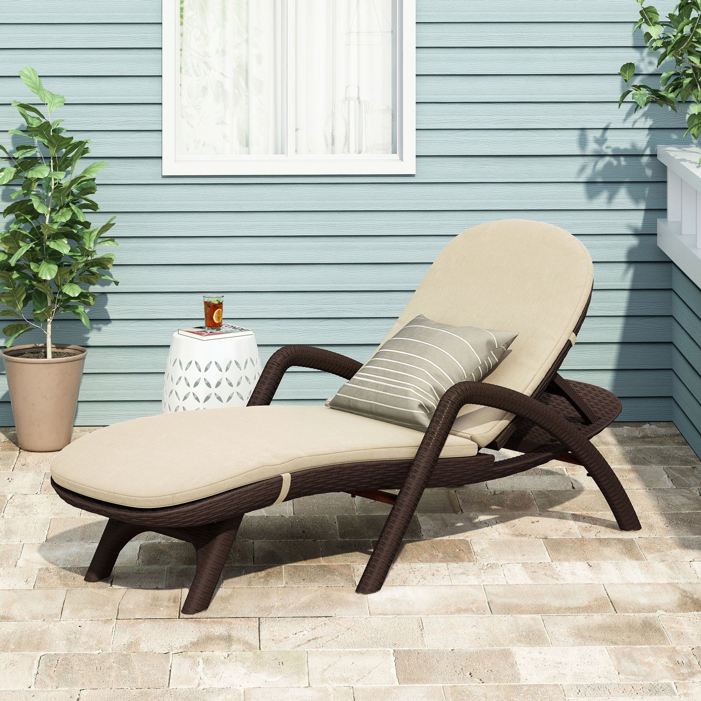 The Mikael - Chaise Lounge With Cushion sits on a stone patio with beige cushions and a striped pillow, next to a small white side table holding a glass of tea; two potted plants and a blue wall are in the background.