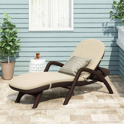 The Mikael - Chaise Lounge With Cushion sits on a stone patio with beige cushions and a striped pillow, next to a small white side table holding a glass of tea; two potted plants and a blue wall are in the background.