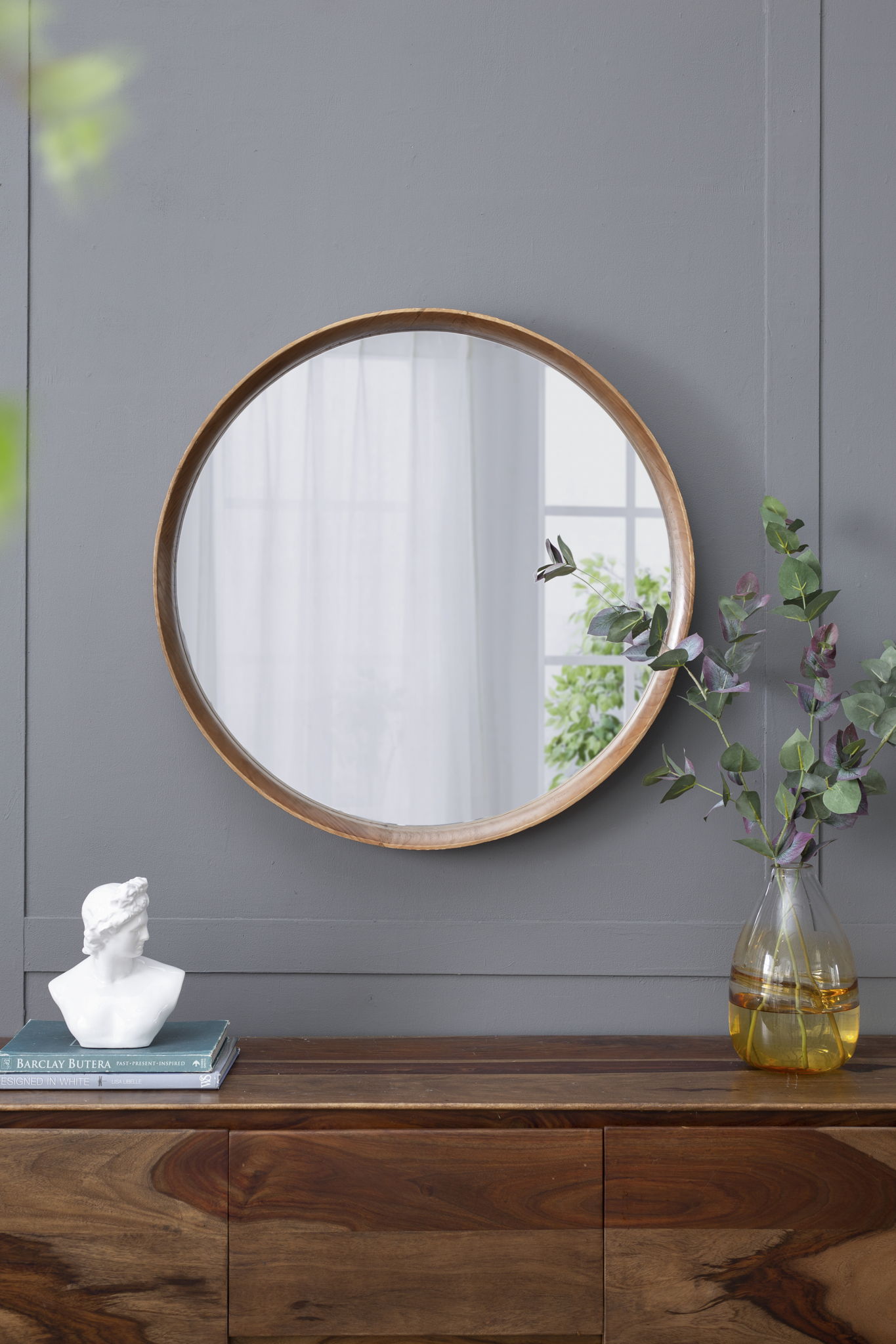 A Round Wood Mirror, Wall Mounted Mirror Home Decor For Bathroom Living Room, hangs on a gray wall above a wooden sideboard with a vase of green branches, a white bust, and books. Natural light streams in from a nearby window.