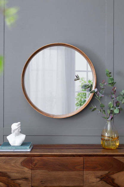 A Round Wood Mirror, Wall Mounted Mirror Home Decor For Bathroom Living Room, hangs on a gray wall above a wooden sideboard with a vase of green branches, a white bust, and books. Natural light streams in from a nearby window.