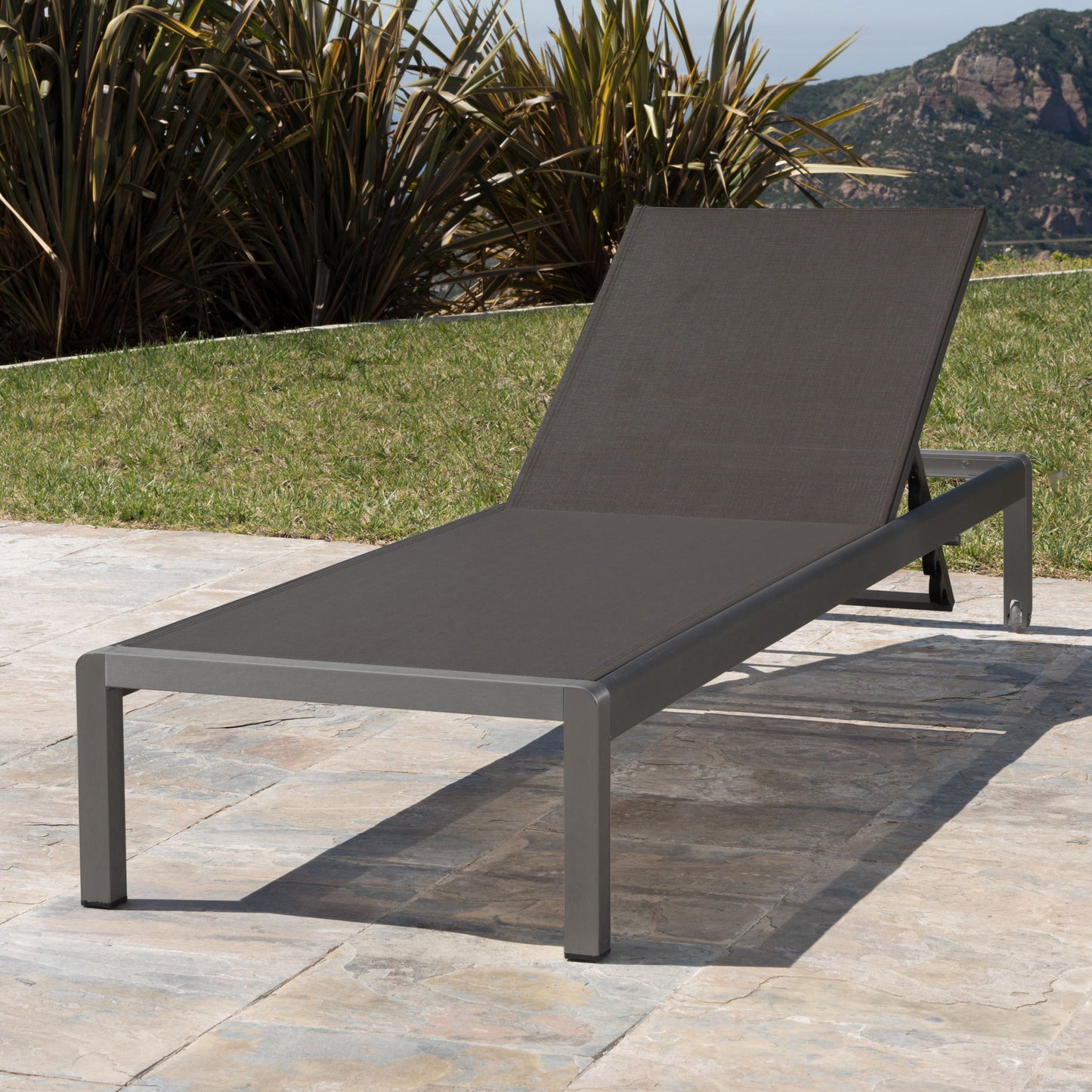 The Outdoor Patio Chaise Lounge With 5 Reclining Positions, featuring a dark metal frame and mesh fabric seat, is placed on a stone patio surrounded by green grass, tall plants, and mountain views in the background.