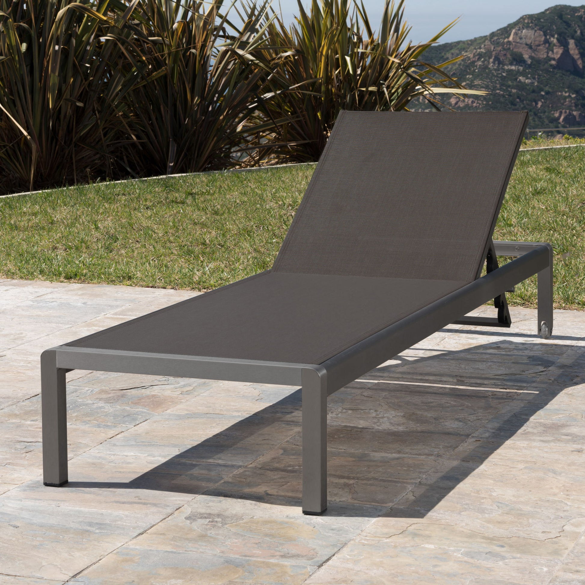 The Outdoor Patio Chaise Lounge With 5 Reclining Positions, featuring a dark metal frame and mesh fabric seat, is placed on a stone patio surrounded by green grass, tall plants, and mountain views in the background.