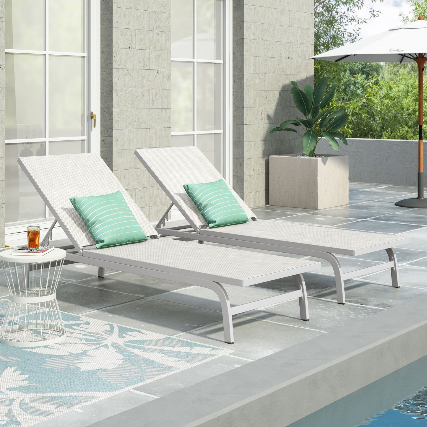 Two Finn Contemporary Chaise Lounges sit poolside with green striped pillows on a tiled patio next to a small side table holding a drink. Plants, an umbrella, and large windows create an inviting outdoor relaxation space.