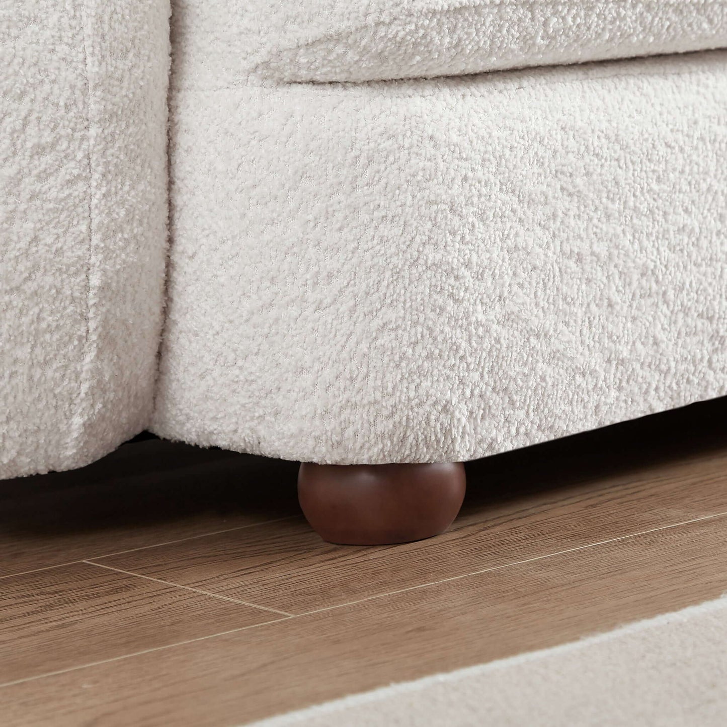 Close-up of the Paton - Comfortable Sofa in light beige with a textured finish and round wooden legs, shown on a wooden floor next to a light-colored rug.