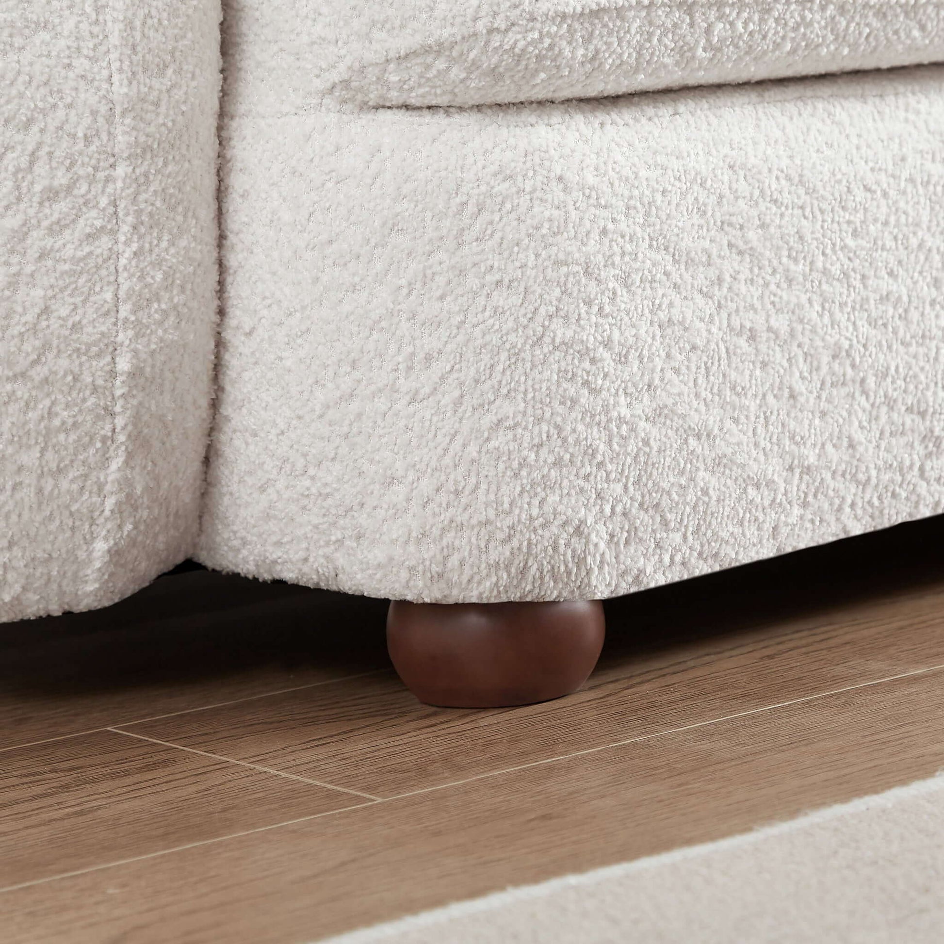 Close-up of the Paton - Comfortable Sofa in light beige with a textured finish and round wooden legs, shown on a wooden floor next to a light-colored rug.