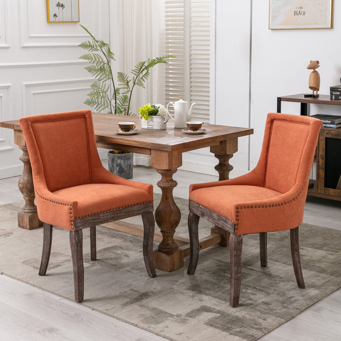A wooden dining table with tea set, two Ultra Side Dining Chairs featuring thick fabric, neutral solid wood legs, and bronze nail heads, a potted plant, and minimalist decor in a bright modern room.