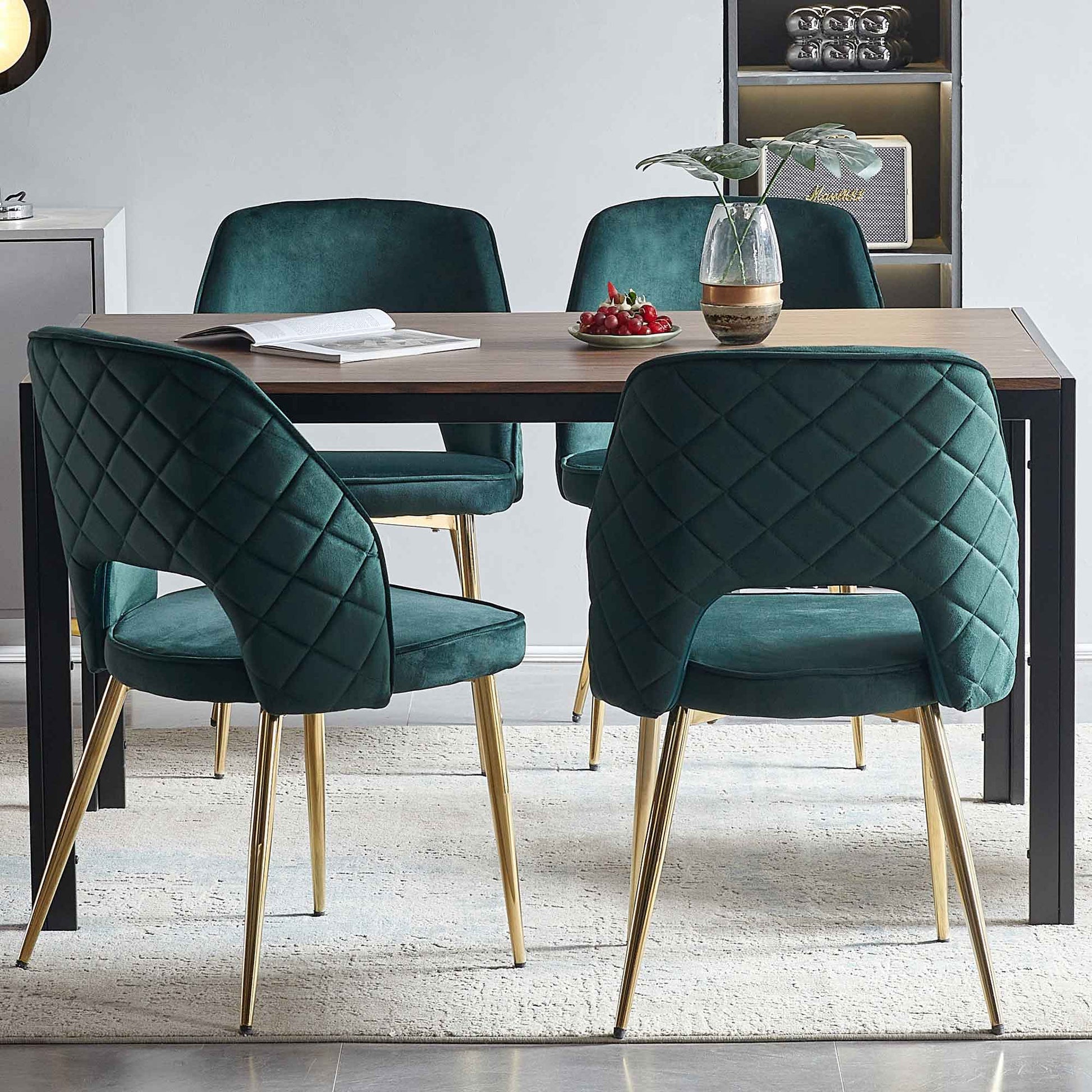 Four Velvet Dining Chairs With Metal Legs And Hollow Back surround a rectangular wooden table topped with an open book, small plant vase, and plate of red berries, all set on a light rug in a modern dining room.