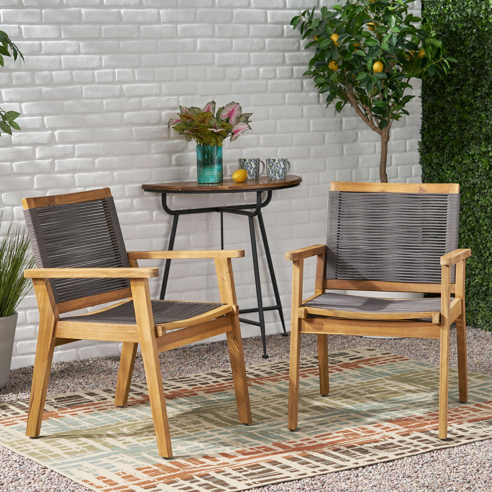 Two Outdoor Dining Chairs (Set of 2) in Dark Gray are arranged on a patterned rug outside, with a small table featuring a vase, cups, and a citrus tree placed against a white brick wall.