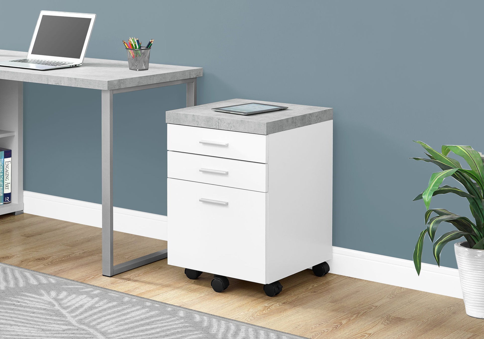 The File Cabinet Rolling Mobile Storage Drawers Printer Stand For Office in white and gray sits by a matching desk with a laptop and a tablet on top, against a blue wall, on a wooden floor with a plant and gray-patterned rug.