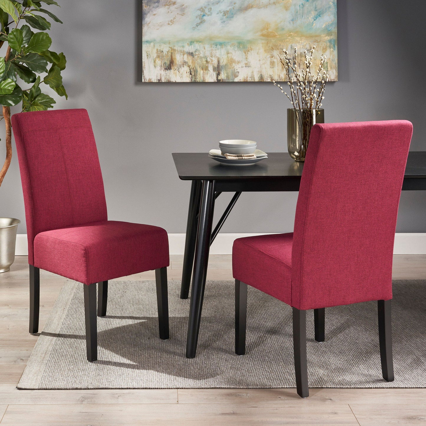 Two Pertica Dining Upholstered Armless Chairs in red with black legs sit around a black dining table set with plates and cups. The modern room features a gray rug, a potted plant, and a framed abstract painting.