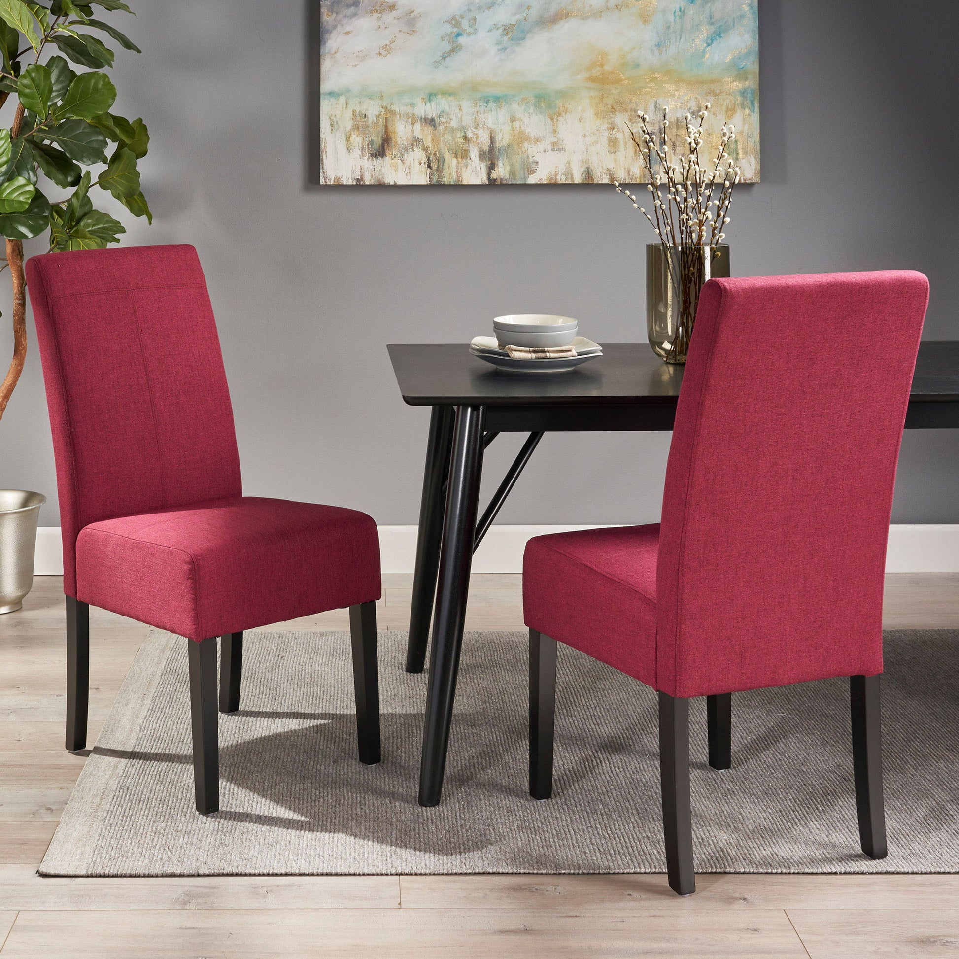 Two Pertica Dining Upholstered Armless Chairs in red with black legs sit around a black dining table set with plates and cups. The modern room features a gray rug, a potted plant, and a framed abstract painting.