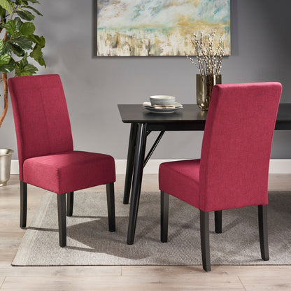 Two Pertica Dining Upholstered Armless Chairs in red with black legs sit around a black dining table set with plates and cups. The modern room features a gray rug, a potted plant, and a framed abstract painting.