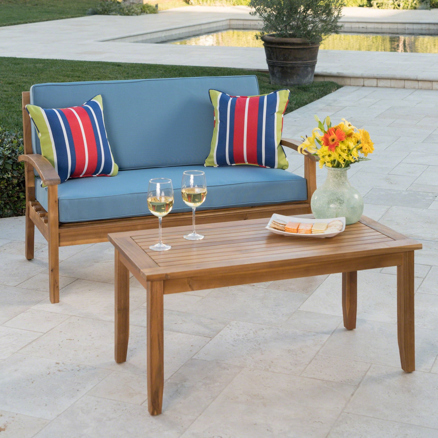 Enjoy a relaxing patio scene by the pool with the Outdoor Chat Set With Coffee Table And Loveseat, featuring blue cushions, striped pillows, white wine glasses, a plate with a napkin, and a vase of yellow flowers.