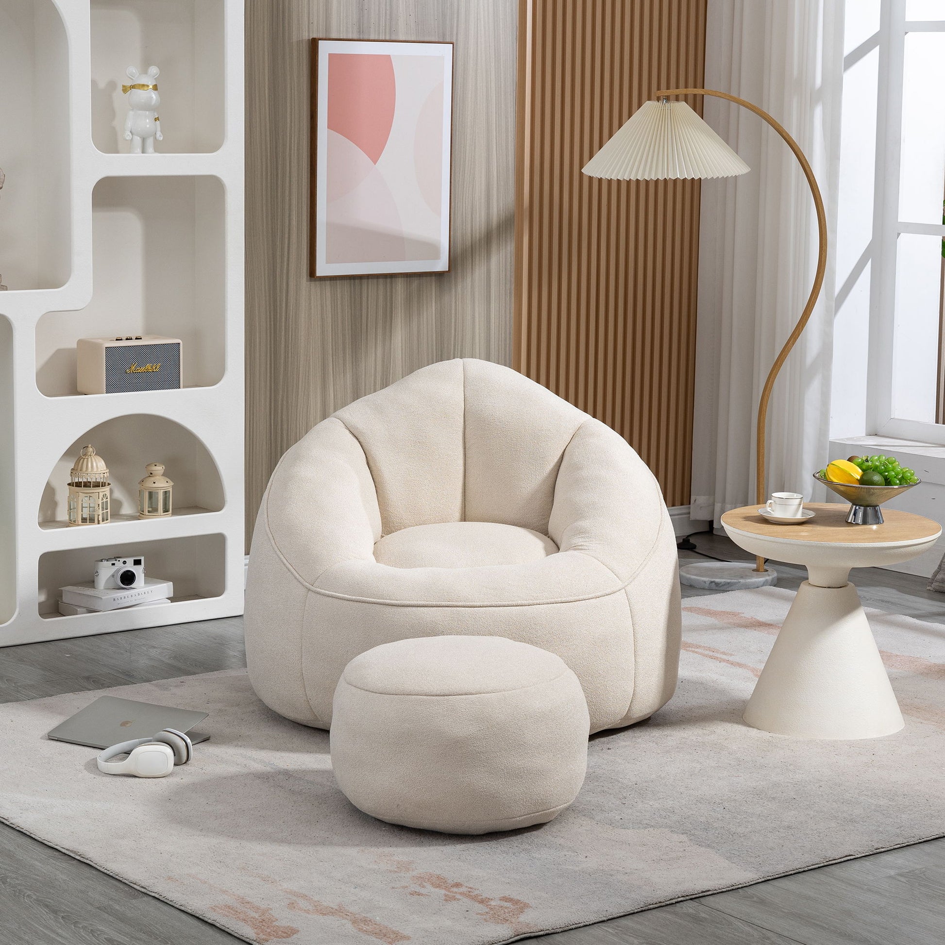 The Bedding Bean Bag Sofa Chair High Pressure Foam with Footrest sits on a light rug in a modern living room, next to a small round table with fruit, a lamp, shelves with decor, headphones, and a window with sheer curtains.