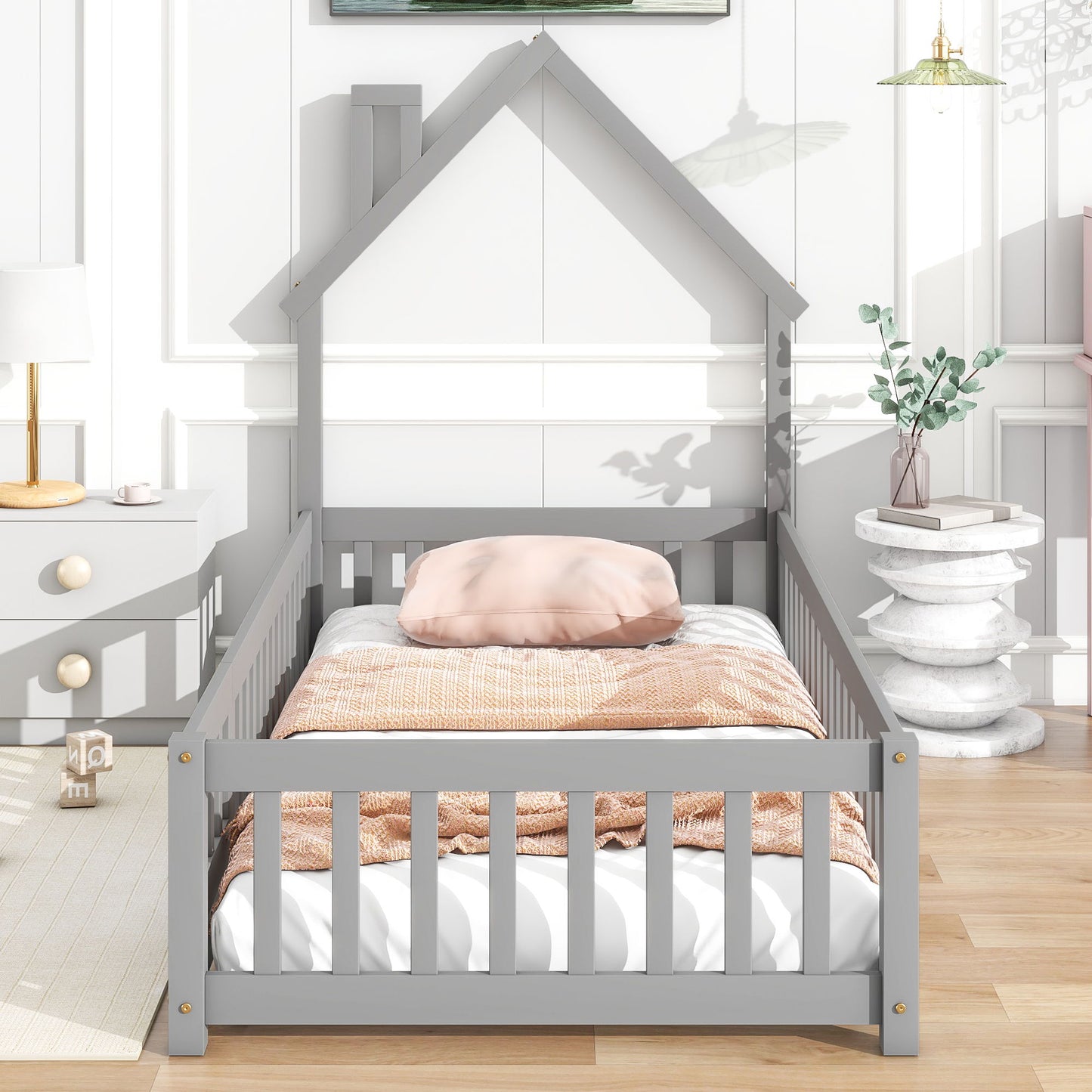 The House-Shaped Headboard Floor Bed With Fence, featuring a pale pink pillow and peach blanket, is set in a bright kids' room with plants, light wood floors, and modern decor.