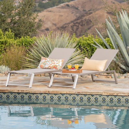 Two Myers Modern Chaise Lounge chairs with colorful pillows sit by a pool, featuring a tray with two glasses of orange juice and fruit. The scene includes succulent plants and hills in the background.