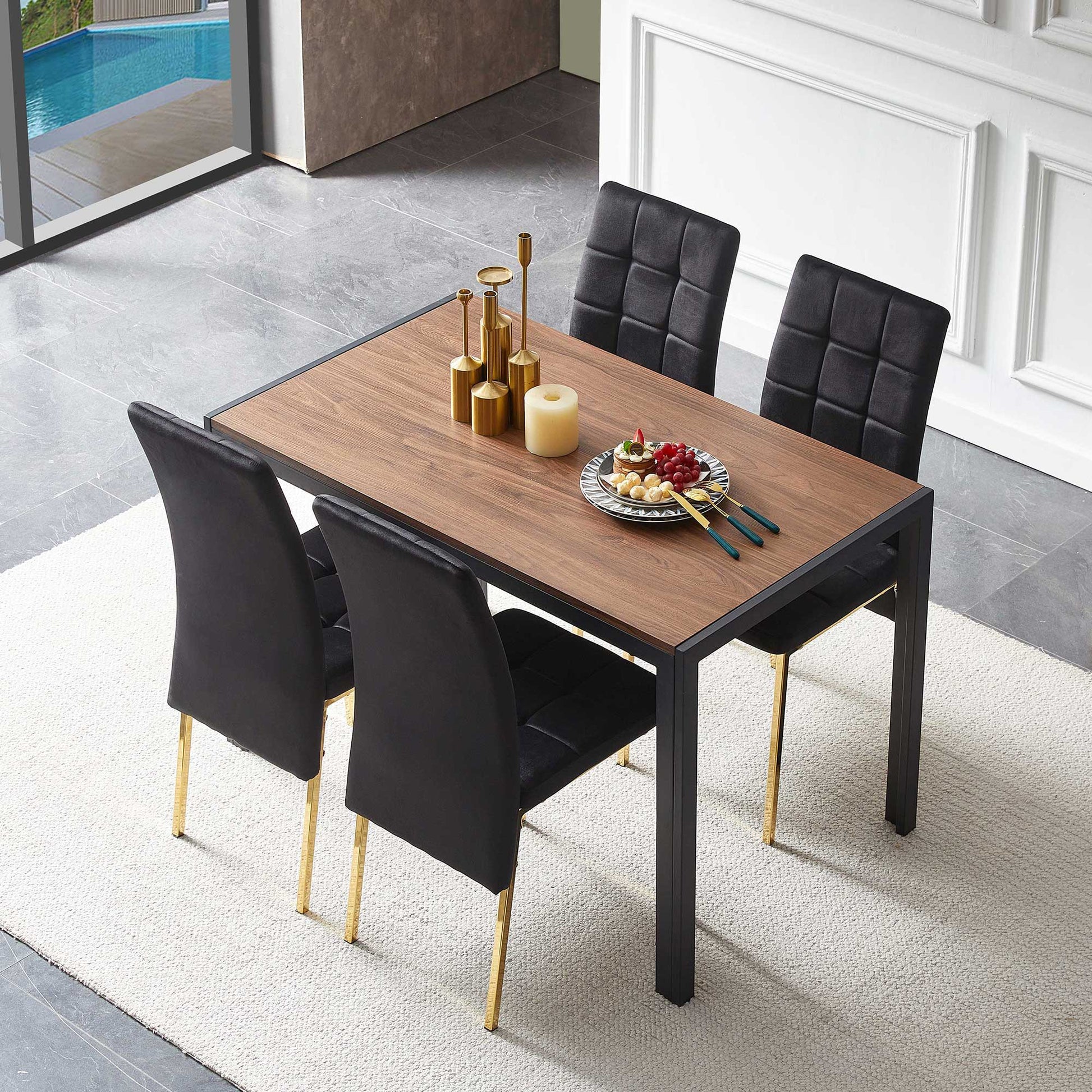 The 5 Pieces Dining Set features a creative dining table and velvet high back Nordic chairs with golden legs, set for four. Displayed on a light rug in a bright room, the table is styled with candles, gold vases, fruit, and cutlery.