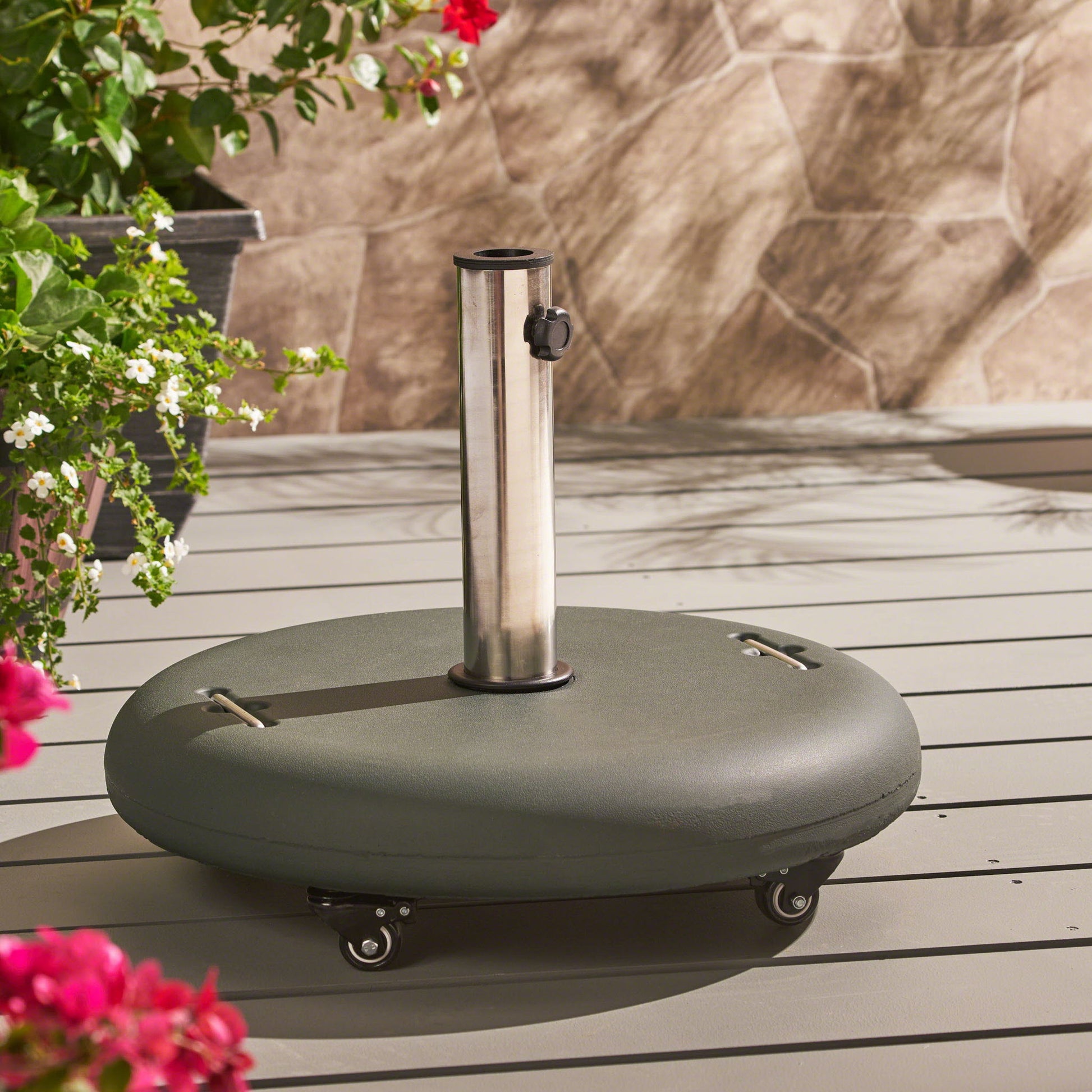 The Wheelie - Round Umbrella Base With Stainless Steel Holder sits on a patio deck with wheels, surrounded by green plants and pink flowers, and backed by a stone wall.