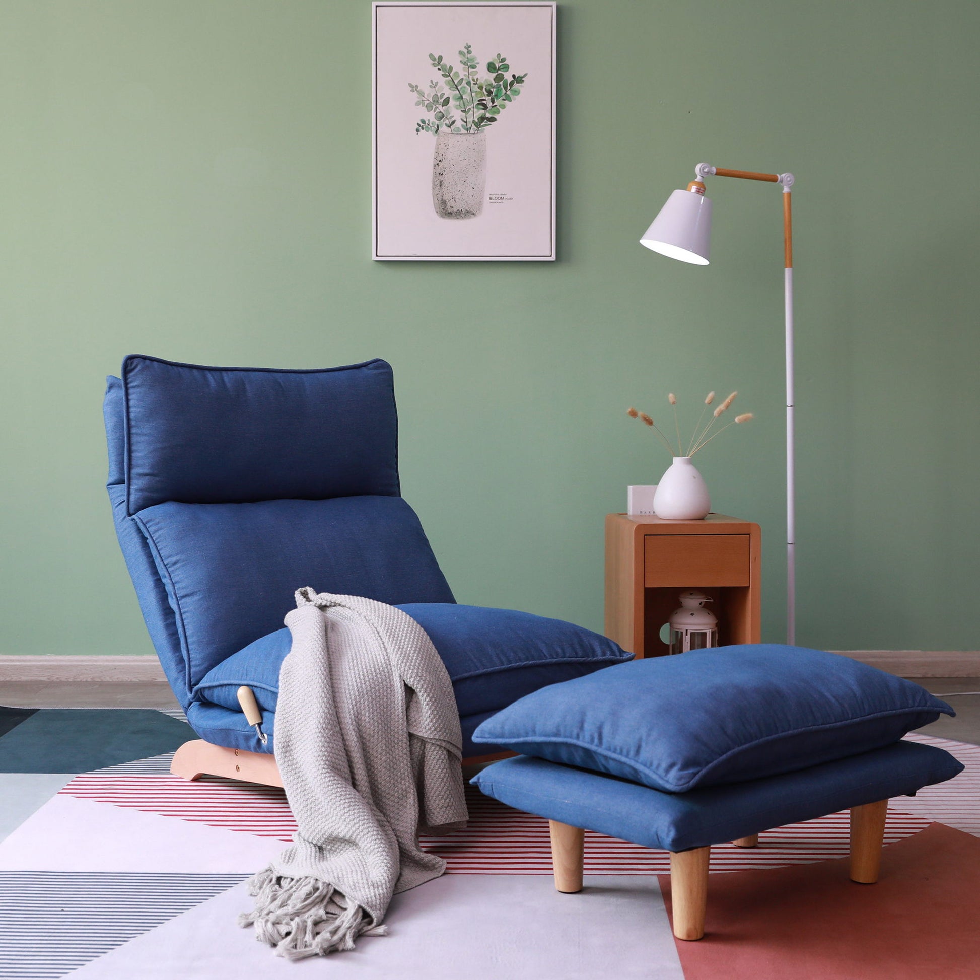 A Single Lazy Sofa Chair Leisure Foldable Reclining Chair in blue with a matching footrest sits on a geometric rug, draped with a gray blanket. A wooden side table and white floor lamp stand nearby against a green wall with framed plant art.