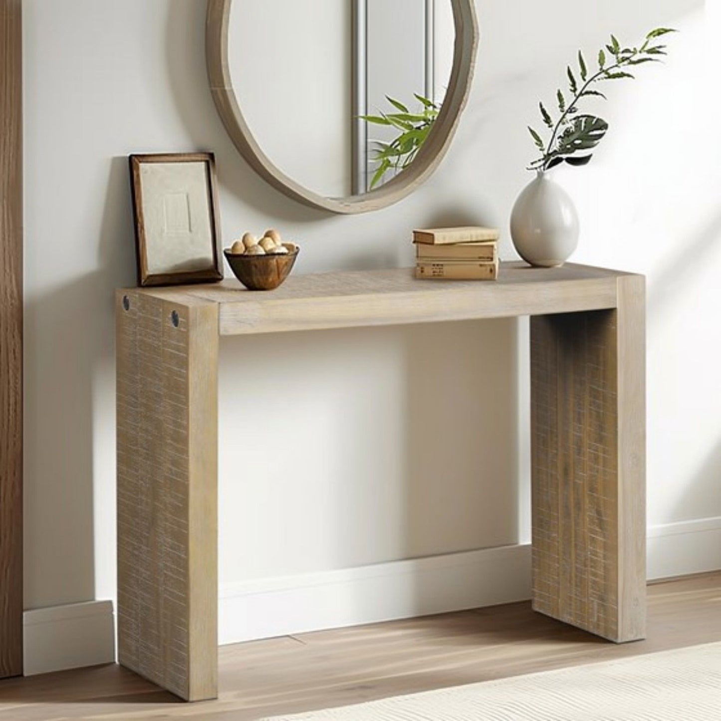 The Modern Design Console Table sits against a neutral wall with a round mirror, framed picture, bowl of nuts, vase of green branches, and two stacked books on top. Light wood flooring completes the scene.