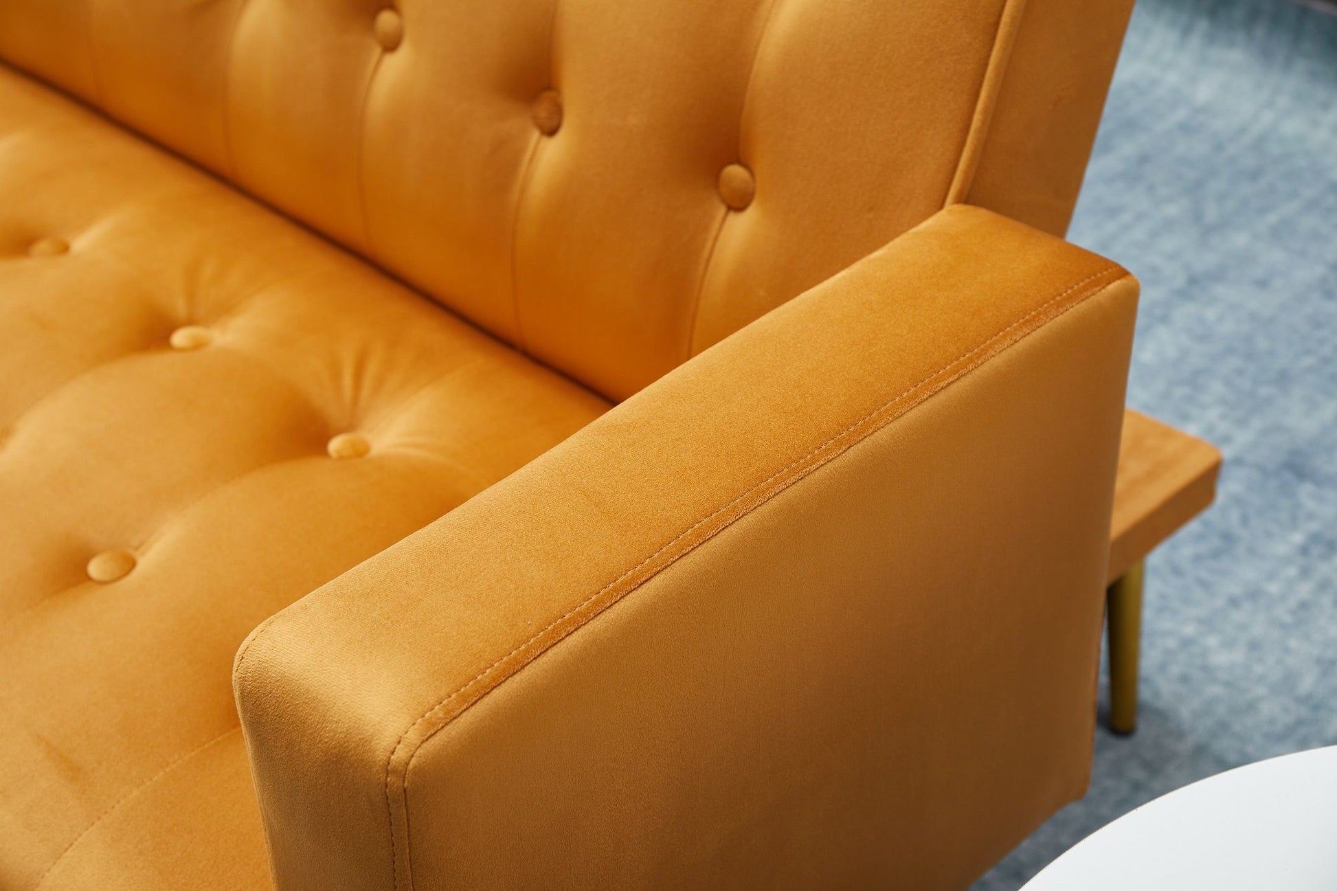 A close-up of the Comfort Compact Design Sofa Bed in mustard yellow velvet, showing its tufted armrest and seat cushion, with a light blue carpet and a partially visible white circular table.