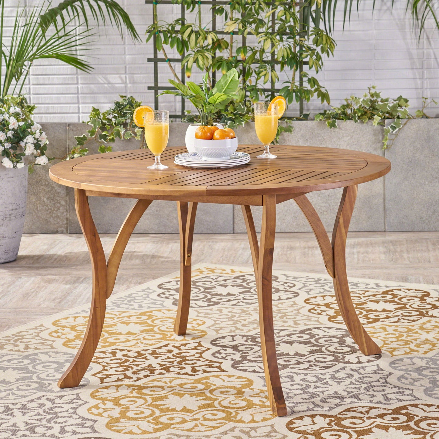 The Hermosa Outdoor Acacia Wood Table with a circular design is set outdoors on a patterned rug, featuring three glasses of orange juice, a bowl of oranges, and stacked white plates, surrounded by green plants.