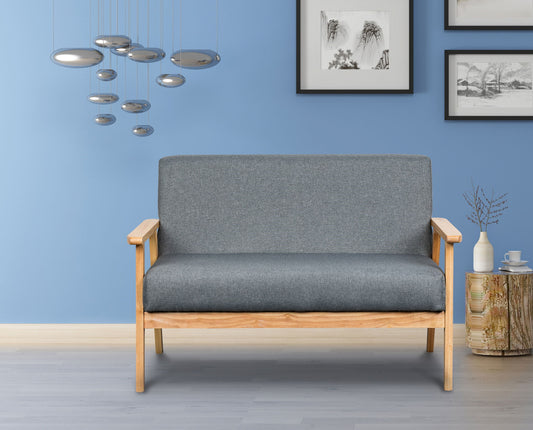 A modern living room with the Bahamas - Loveseat in light wood and gray cushions, blue walls, silver pendant lights, framed art, a wicker side table, and decorative accents like a vase and branches.