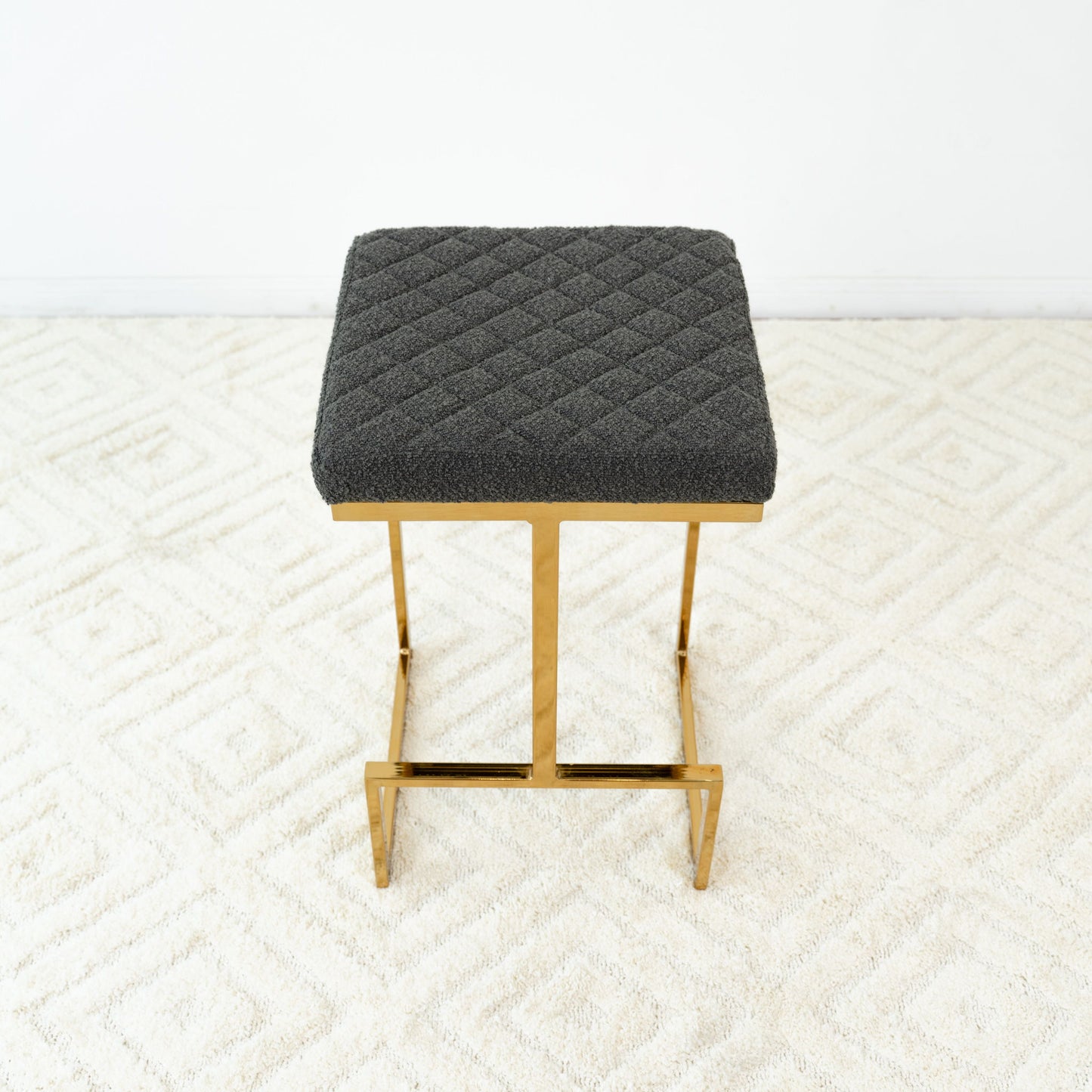 The Joel Mid Century Modern Luxury Upholstered Stool features a quilted dark gray cushion and gold metal legs, shown on a light patterned carpet with a plain white wall backdrop.