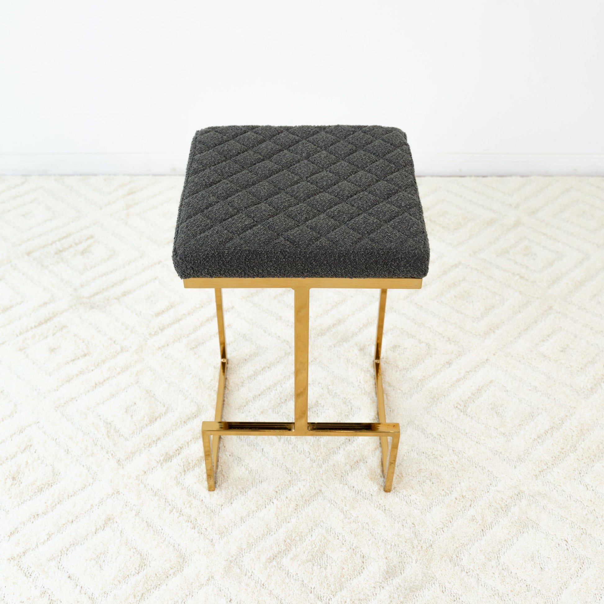 The Joel Mid Century Modern Luxury Upholstered Stool features a quilted dark gray cushion and gold metal legs, shown on a light patterned carpet with a plain white wall backdrop.