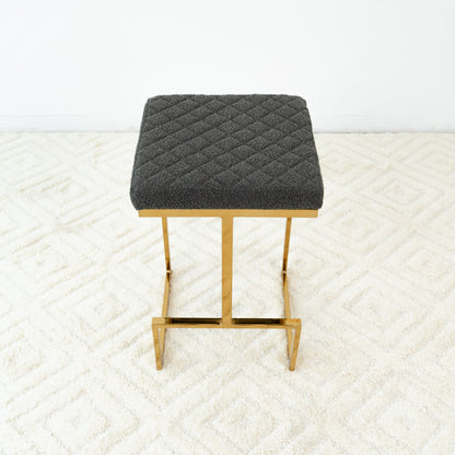 The Joel Mid Century Modern Luxury Upholstered Stool features a quilted dark gray cushion and gold metal legs, shown on a light patterned carpet with a plain white wall backdrop.