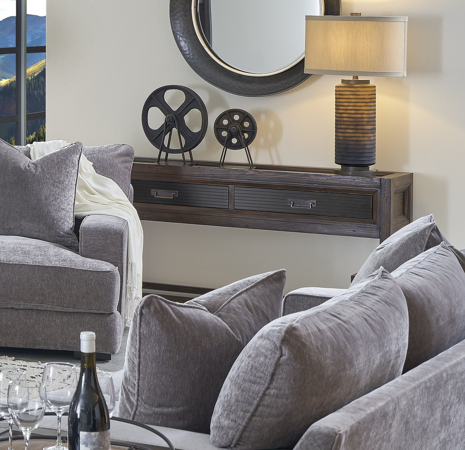 A cozy living room with two gray sofas, a Couch.com Branson 2 Drawer Sofa Table in dark brown wood, metal decor pieces, a round mirror, table lamp, throw blanket on the sofa, and a wine bottle on the glass coffee table.