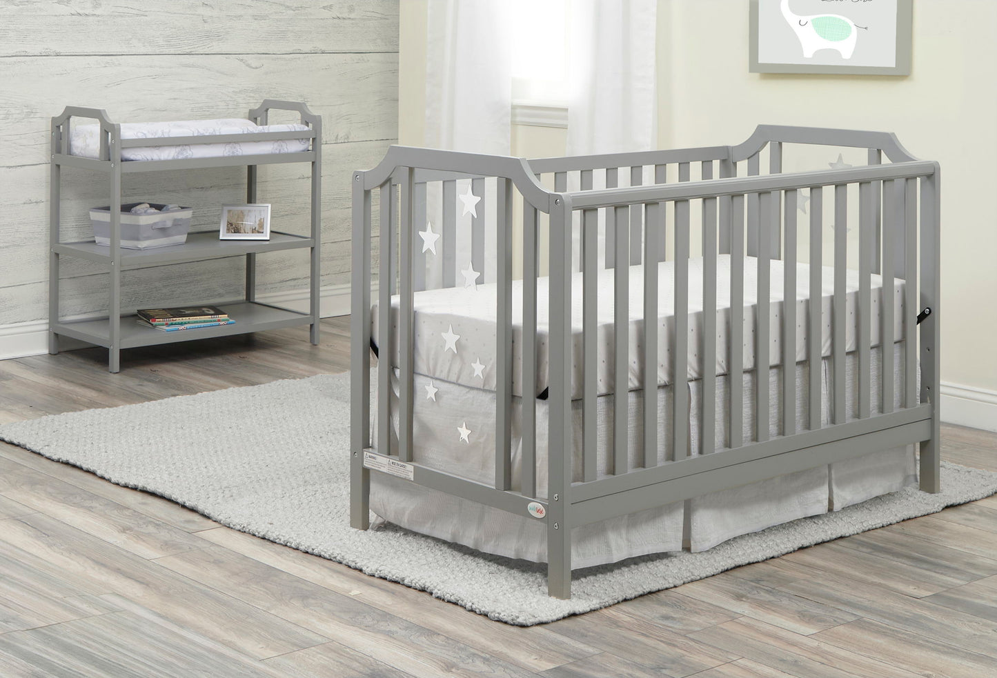 A gray baby crib with star-patterned bedding sits on a gray rug in the nursery. The Celeste Changing Table with shelves and storage baskets is placed against the wall, by a window with sheer curtains.