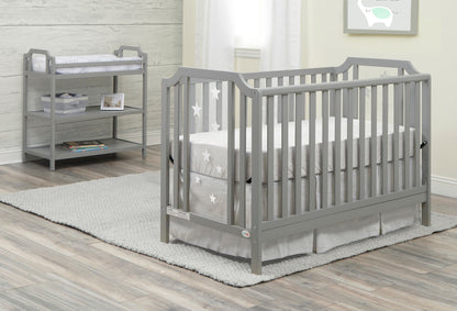 A gray baby crib with star-patterned bedding sits on a gray rug in the nursery. The Celeste Changing Table with shelves and storage baskets is placed against the wall, by a window with sheer curtains.