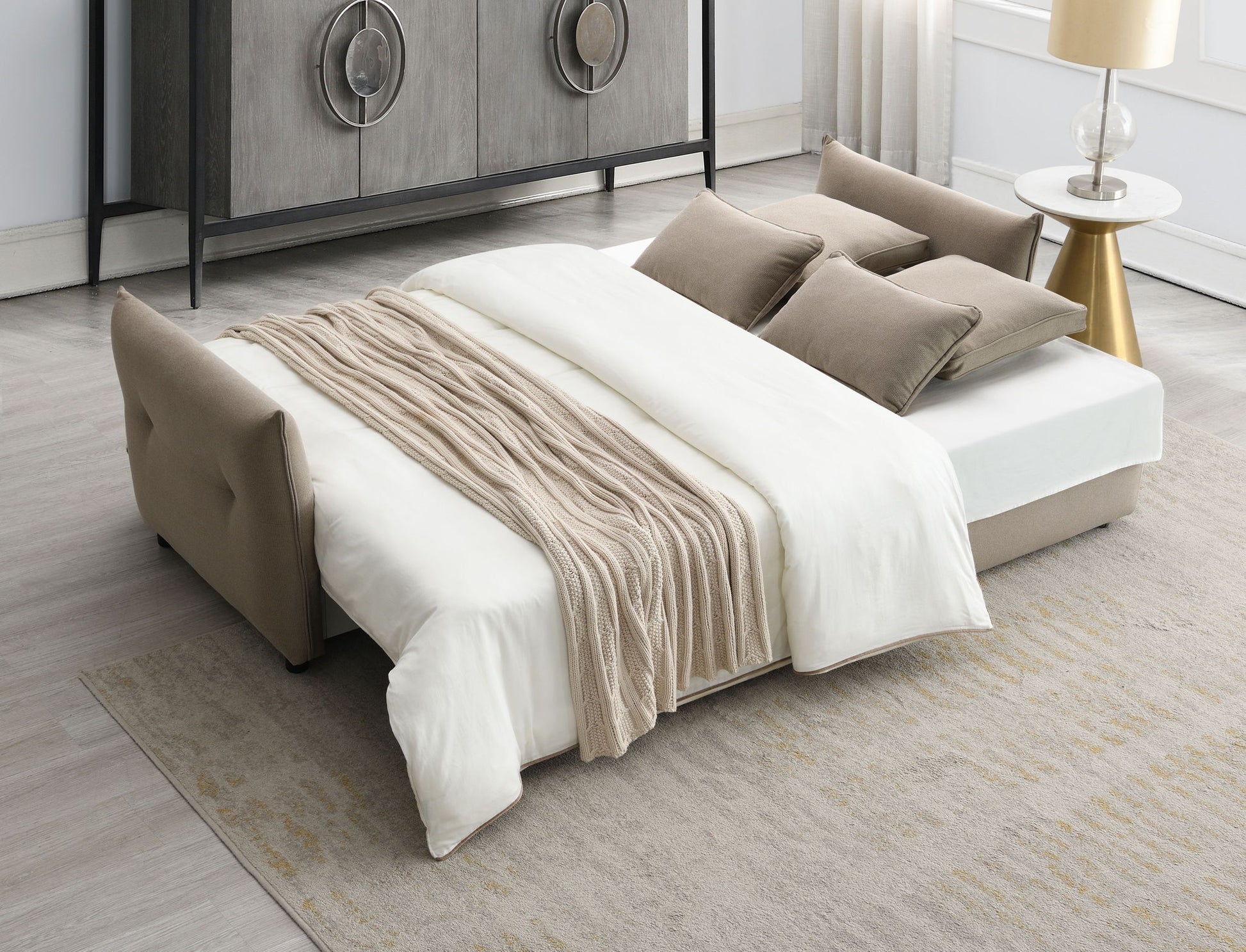 The Irina Pull Out Sleep With Storage, featuring white sheets, a beige knit blanket, and matching pillows, is arranged in a modern living room with a gray rug, gold side table, and dresser in the background.