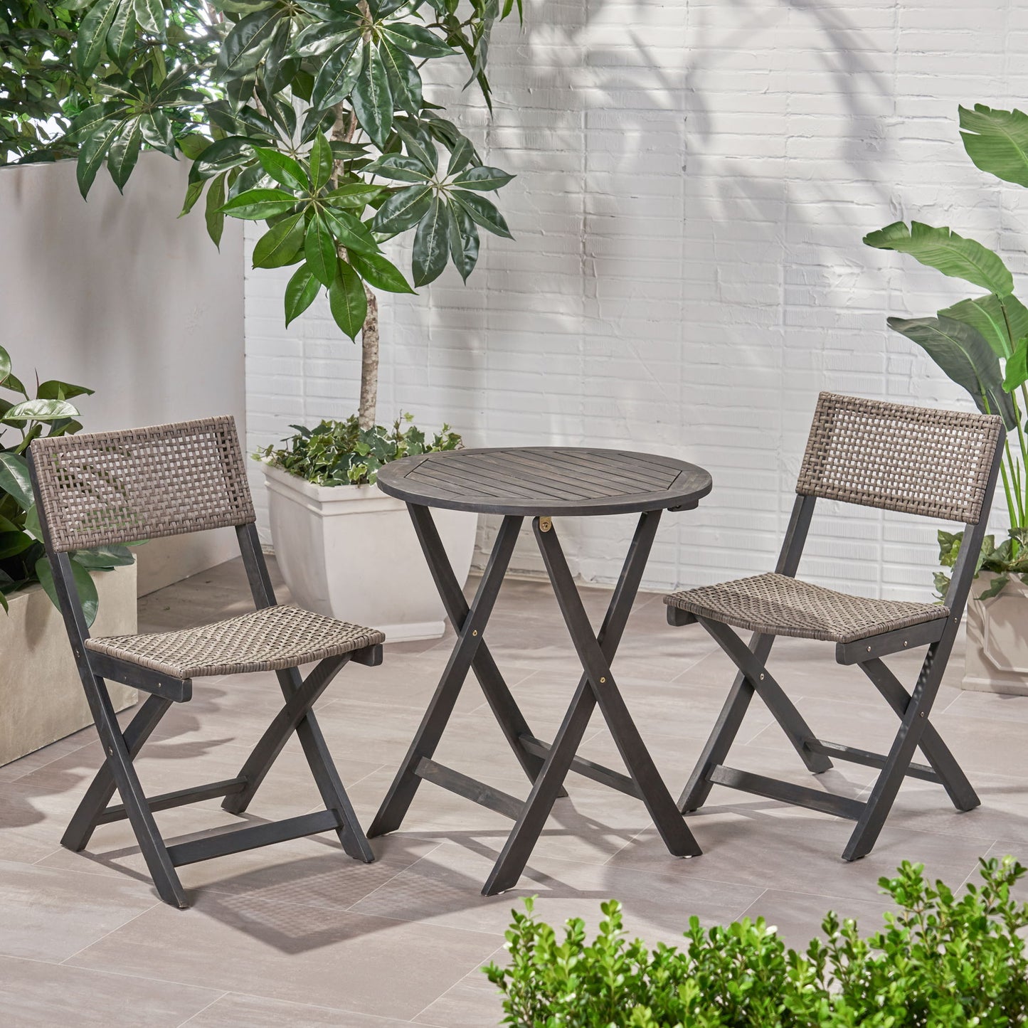 The Evergreen Outdoor Dining Set, featuring a round wooden table and two matching chairs, is arranged on a tiled patio surrounded by potted green plants near a white brick wall.