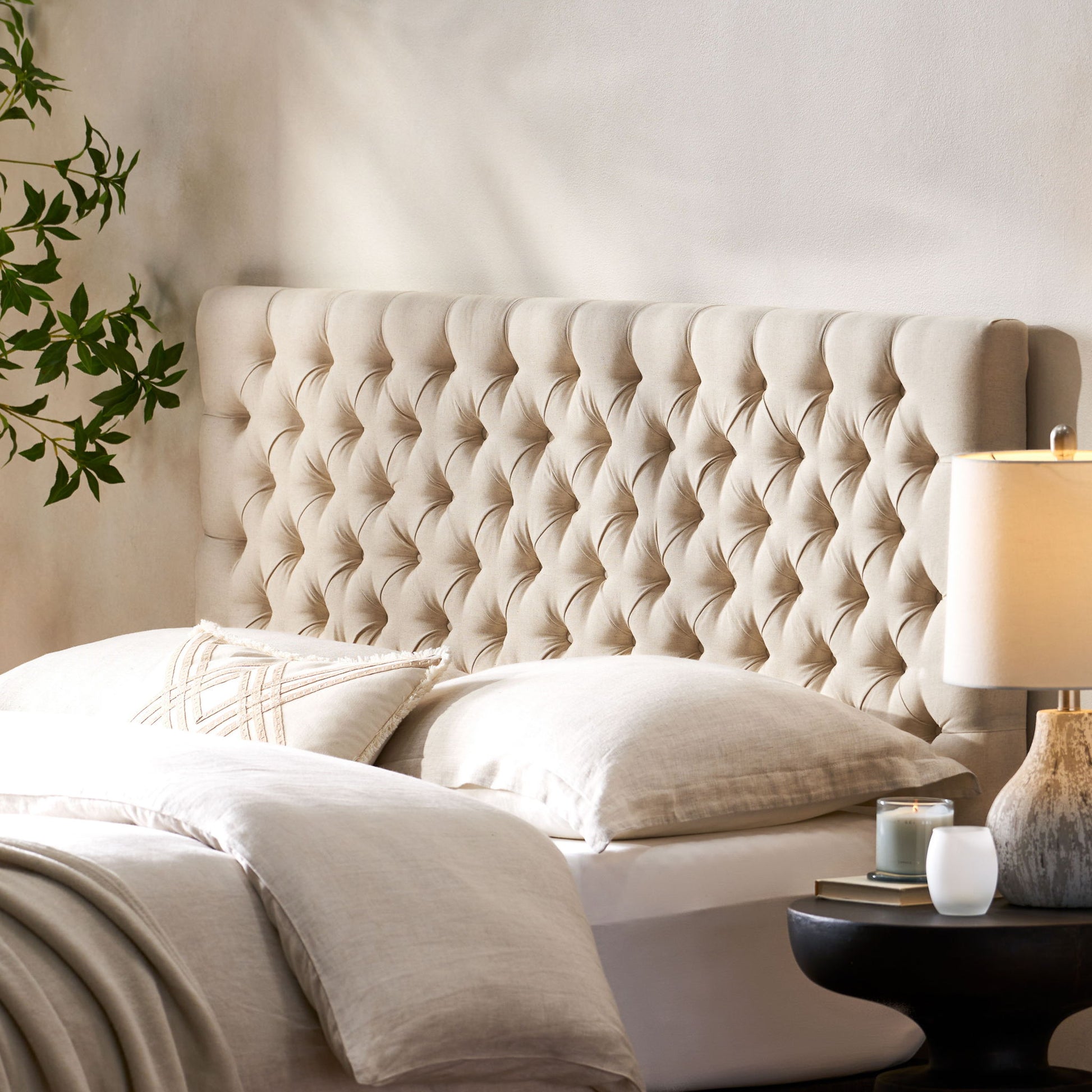 A cozy bedroom featuring the Annecy Contemporary Upholstered Headboard in beige, cream bedding, a round black nightstand with a lit candle and lamp, plus a leafy plant against a light-colored wall.